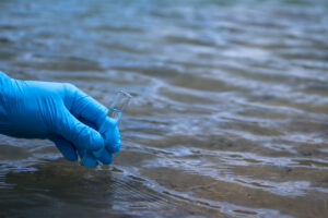 A man's hand in a blue glove takes a close-up of water into a test tube to measure water pollution. Background